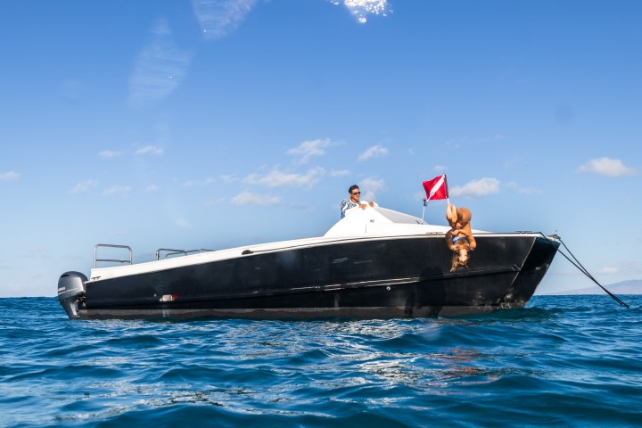 Person dives off black boat in blue sea under a clear sky.
