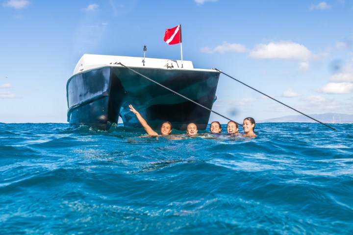 Five people swimming near a boat with a red flag in open water under a clear blue sky.