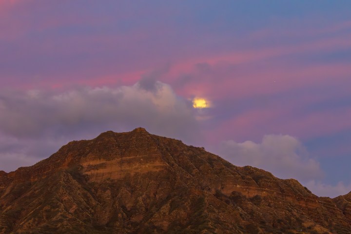 Mountain silhouette at dusk with colorful sky and moon partially hidden by clouds.