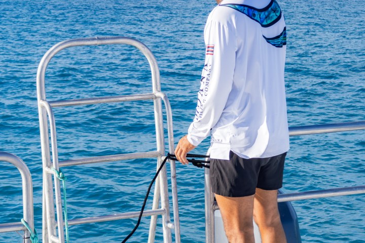 Person on boat deck in shorts and long-sleeve shirt, holding a hose facing the ocean.