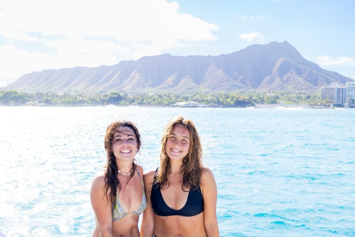 Two women in swimsuits smiling with ocean and mountain in background.