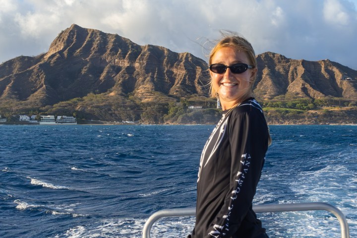 Woman in sunglasses on a boat with mountains and ocean in the background.