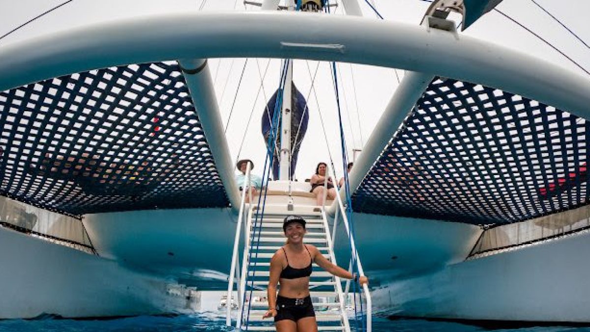 Person climbing onto a catamaran with people lounging on deck, seen from water level.