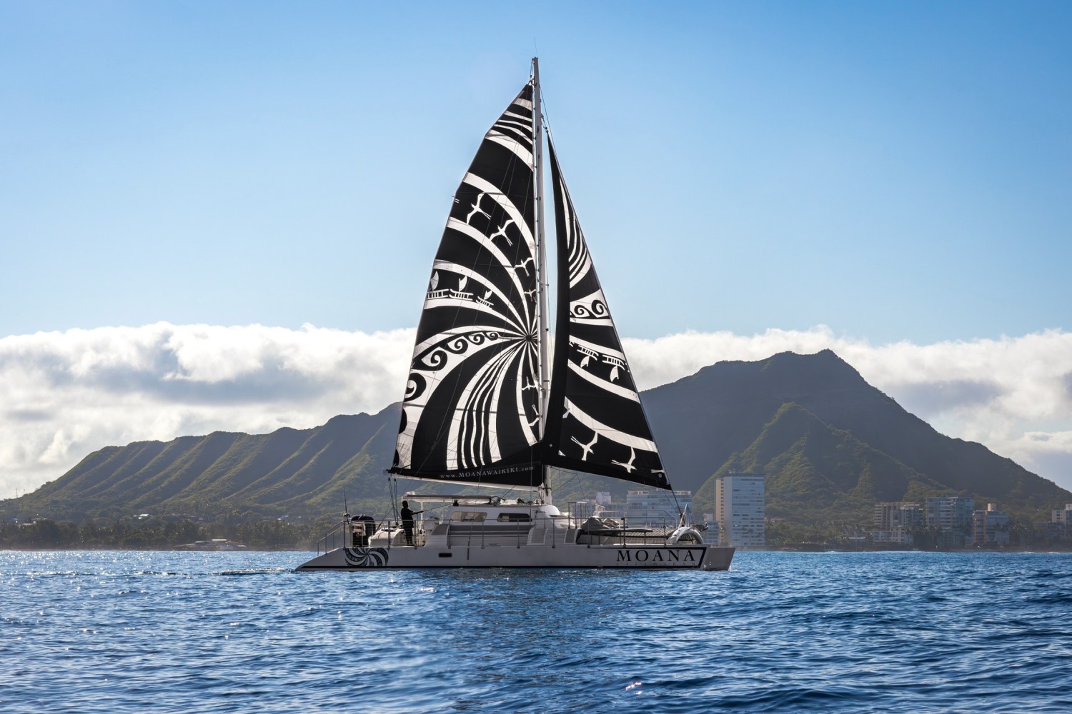 Sailboat with tribal-patterned sail on ocean with mountain and cloudy sky backdrop.